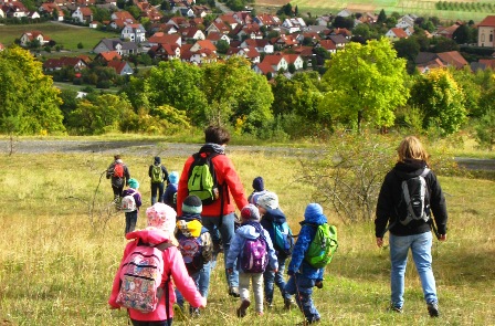 Kinder und Erwachsene mit Rucksack wandern in ein Dorf, Foto: DWV