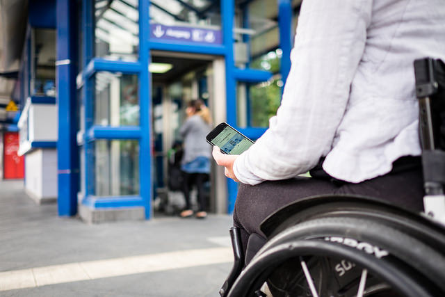 Junger Mensch im Rollstuhl checkt ihr Handy vor einem Aufzug im öffentlichen Nahverkehr